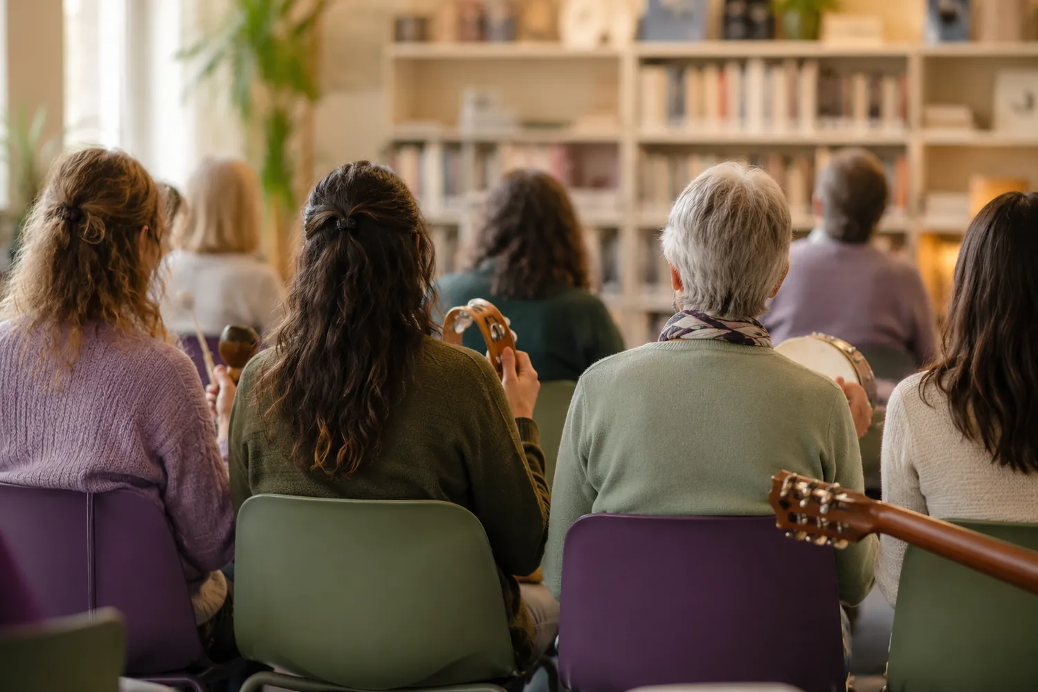 Rows of adults seated in a school library workshop, seen from behind, holding tambourines, maracas, and a hand drum, with a guitar neck in the foreground