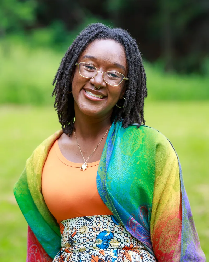 Anaya Baptiste smiling in a bright rainbow shawl, standing in a sunny green field