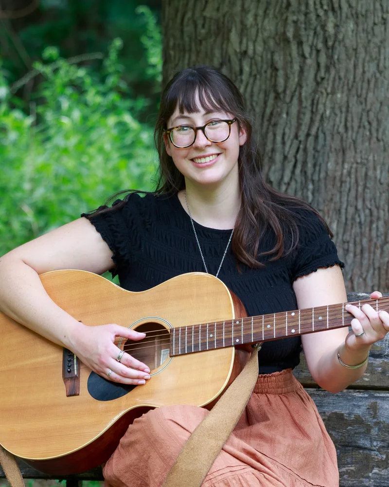 Hannah Baker smiling, seated on a wooden bench outdoors and playing an acoustic guitar