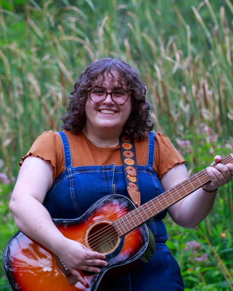 Lauren Tropper smiling and playing an acoustic guitar among tall grasses